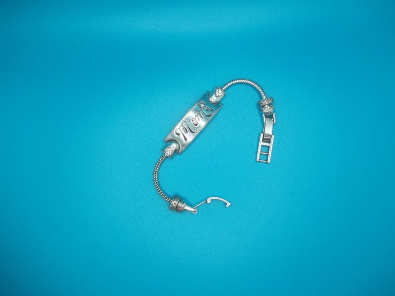 May include: A silver bracelet featuring the word "Mom" engraved on a rectangular charm. The bracelet has a flexible, coiled design with decorative beads and a clasp. The background is a solid teal colour.
