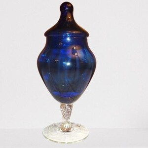 May include: A cobalt blue glass jar with a lid, resting on a clear, twisted stem and a round base. The jar has a bulbous body and a rounded lid. The glass has a reflective, glossy finish.