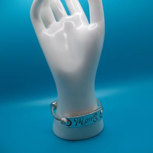 May include: A silver bracelet inscribed with the word "Mom" in cursive is presented on a white hand-shaped jewellery holder against a blue backdrop. The bracelet features a decorative design, a thoughtful gift.
