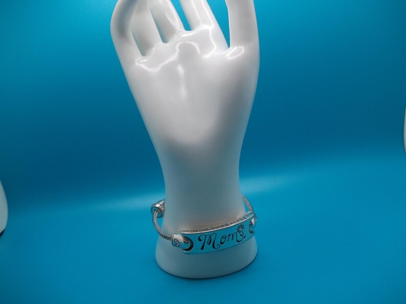 May include: A silver bracelet inscribed with the word "Mom" in cursive is presented on a white hand-shaped jewellery holder against a blue backdrop. The bracelet features a decorative design, a thoughtful gift.