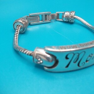 May include: Silver bracelet with the word "Mom" cut out on a curved plate. The bracelet has a flexible, snake-like chain and decorative end pieces. The background is a solid blue.