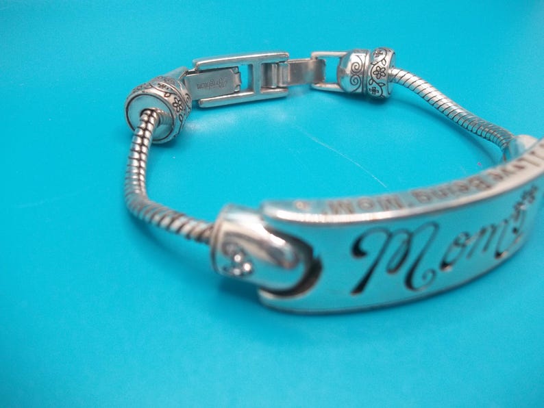 May include: Silver bracelet with the word "Mom" cut out on a curved plate. The bracelet has a flexible, snake-like chain and decorative end pieces. The background is a solid blue.