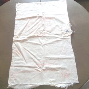 May include: White cotton tablecloth with a blue and white embroidered flower design in the upper right corner. The tablecloth has a frayed edge and is slightly wrinkled.