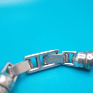 May include: Close-up of a silver bracelet clasp against a bright blue background. The clasp is rectangular with the brand name engraved on it. The bracelet has a snake chain design.