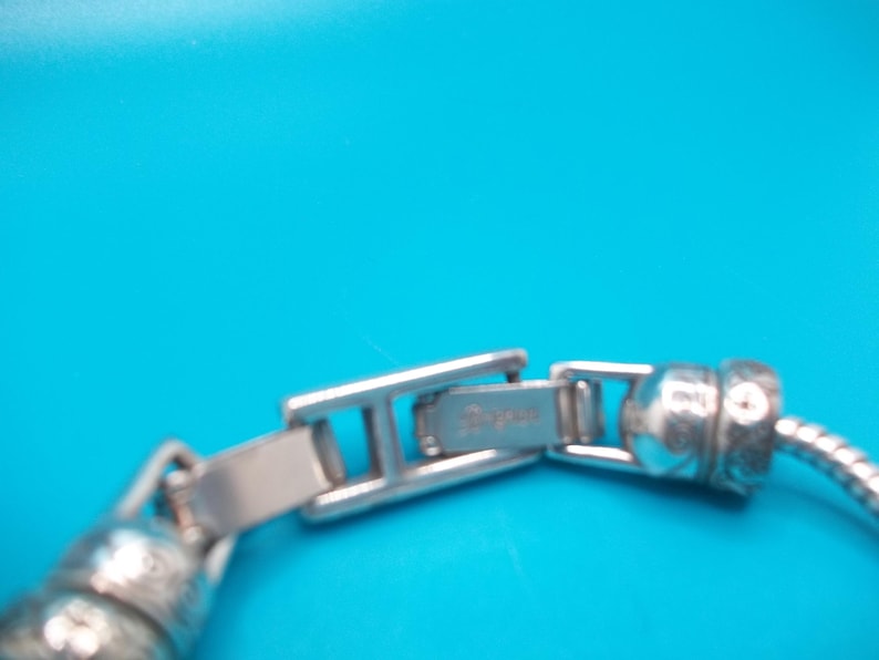 May include: Close-up of a silver bracelet clasp against a bright blue background. The clasp is rectangular with the brand name engraved on it. The bracelet has a snake chain design.