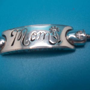 May include: Silver charm bracelet bead with the word "Mom" in cursive script. The bead has a floral design and is connected to a silver chain. The background is a solid blue.
