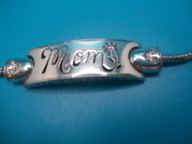May include: Silver charm bracelet bead with the word "Mom" in cursive script. The bead has a floral design and is connected to a silver chain. The background is a solid blue.