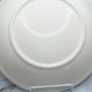 May include: A white ceramic plate, viewed from the underside, showcasing a smooth, glossy surface. The plate has a slightly raised rim and a central, recessed area. The design is simple and elegant, suitable for various culinary presentations.