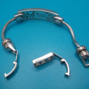 May include: Silver bracelet with the inscription "I love my family" and a decorative floral design. The bracelet is unclasped, showing the clasp mechanism. The background is a solid blue.