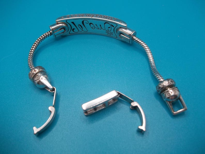 May include: Silver bracelet with the inscription "I love my family" and a decorative floral design. The bracelet is unclasped, showing the clasp mechanism. The background is a solid blue.