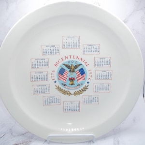 May include: A white ceramic plate with a calendar design commemorating the Bicentennial of 1776. The plate features the American flag, an eagle, and the words "Bicentennial 1776-1976". Calendars for each month are printed around the central design.