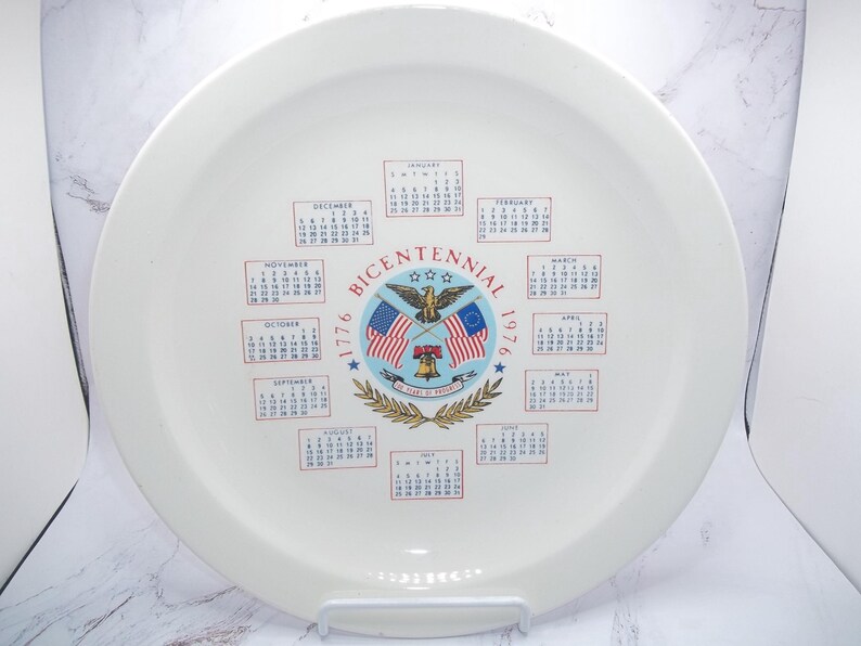 May include: A white ceramic plate with a calendar design commemorating the Bicentennial of 1776. The plate features the American flag, an eagle, and the words "Bicentennial 1776-1976". Calendars for each month are printed around the central design.