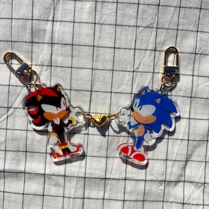 May include: A pair of keychains featuring the characters Sonic the Hedgehog and Shadow the Hedgehog. Sonic is blue with green eyes and Shadow is red with black eyes. The keychains are connected by a gold chain with a heart charm.