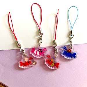 May include: Four clear acrylic charms featuring cartoon shrimp designs. Each charm has a different coloured shrimp: red, pink, and blue. Each shrimp has a different hat and accessory. The charms are attached to a silver clasp and a coloured lanyard.