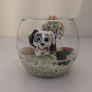 Peut inclure: Un terrarium en verre transparent avec une figurine miniature de chien dalmatien, un petit arbre et d'autres éléments décoratifs. La base est remplie de petits galets de couleur claire. Le chien a des taches noires et un collier rouge.