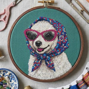 May include: A cross-stitch embroidery hoop featuring a dog wearing sunglasses and a patterned scarf. The image is surrounded by craft supplies, including scissors, thread, and a small dish of pins. A succulent plant in a small pot is also visible.