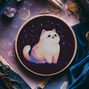 Könnte beinhalten: Ein Kreuzstich-Stickrahmen mit einer pixeligen weißen Katze mit rosa und blauen Akzenten vor einem dunkelblauen Sternenhintergrund. Der Rahmen befindet sich vor einem Hintergrund aus himmlischem Papier, Kristallen und Samt.