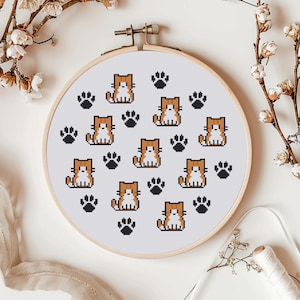 May include: A cross-stitch embroidery hoop with a repeating pattern of pixelated orange and white cats and black paw prints on a white background. The embroidery is set in a wooden hoop.