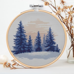 Op de afbeelding: Een borduurring met een wintertafereel van blauwe dennenbomen tegen een witte besneeuwde achtergrond en een lichtblauwe lucht.