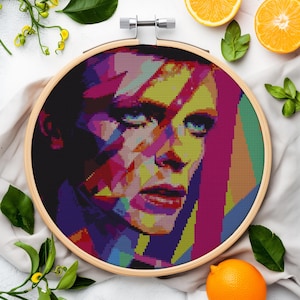 Może przedstawiać: Kolorowe haftowane obrazem twarzy Davida Bowie w drewnianym kółku do haftu. Haftowane jest w pikselowym stylu z jasnymi kolorami, takimi jak różowy, żółty, niebieski i zielony.