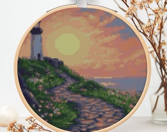 Puede incluir: Una bordado de punto de cruz que representa un faro en un acantilado rocoso con un camino de piedra que conduce hasta él. El cielo es una puesta de sol naranja y rosa vibrante con un gran sol amarillo.