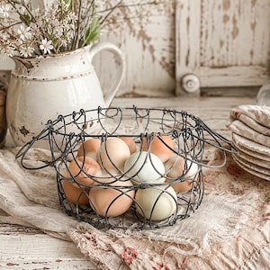 Op de afbeelding: Een ronde draadmand met een donkere metalen afwerking en een decoratieve geschulpte rand, gevuld met een verzameling hele kippeneieren in diverse natuurlijke tinten lichtbruin, beige en bleek blauwgroen, gerangschikt in een open structuur.