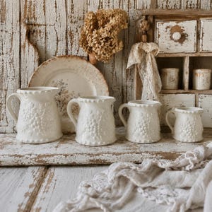 May include: A set of four cream-colored ceramic pitchers of varying sizes, each featuring a raised grape vine relief pattern and a beaded rim. The pitchers are arranged in a row, accompanied by a matching decorative plate and a wooden cabinet.