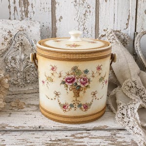 Puede incluir: Un cubo de hielo cilíndrico de cerámica con base de color crema, que presenta un estampado central de ramo de flores en tonos rosa, verde y azul. El recipiente tiene una tapa a juego con un pomo blanco, bordes dorados y asas laterales.
