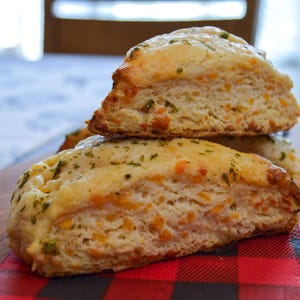 May include: A stack of three golden brown biscuits with a flaky texture and a hint of green herbs. The biscuits are on a red and black checked napkin.