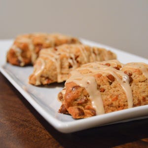 May include: Three baked scones with a light brown glaze, presented on a white rectangular plate. The scones have a golden-brown color with visible nuts and a drizzle of creamy glaze. The plate sits on a dark wooden surface.