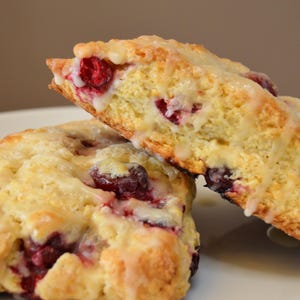 May include: Two triangular scones with a white glaze and red cranberries. The scones are stacked on top of each other on a white plate.