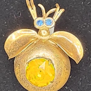 Op de afbeelding: Een gouden broche in de vorm van een insect met blauwe edelsteenogen en een gele edelsteen in het midden.