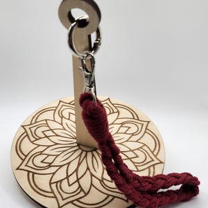 May include: A wooden stand with a mandala design and a red cord attached to a metal ring. The stand is designed to hold a hammock.