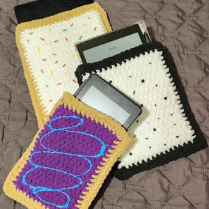 May include: Crocheted tablet sleeves in the shape of pastries. One sleeve is white with colorful sprinkles, another is purple with blue swirls, and the third is white with black dots. Each sleeve has a tan border.