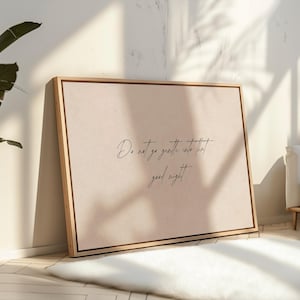 May include: A framed canvas print with the quote "Do not go gentle into that good night" in elegant script. The artwork features a light beige background and a natural wood frame, leaning against a wall. The room is lit by natural light.