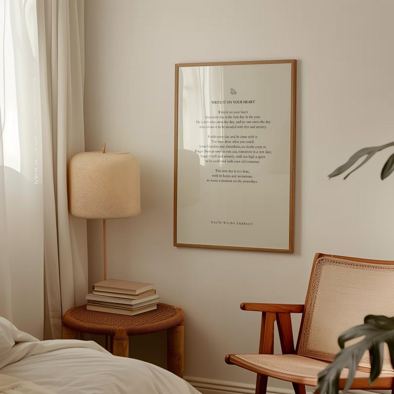 May include: A framed print with a white background and black text. The text reads "Write it on your heart..." and is a quote by Ralph Waldo Emerson. The print is hanging on a light beige wall in a room with a wooden chair, a lamp, and a side table with books.