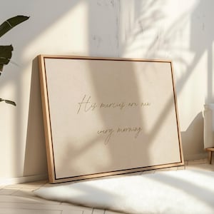 May include: A framed canvas print with a light beige background and the text "His mercies are new every morning" in gold script. The frame is made of light-colored wood. The print is leaning against a wall.