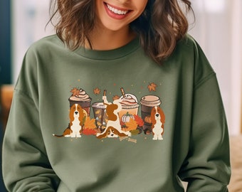 May include: An olive green sweatshirt with a graphic of Cavalier King Charles Spaniels, autumn-themed coffee cups, pumpkins, and leaves. The dogs are in different poses. The sweatshirt has a crew neck.