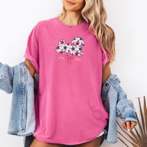 May include: A vibrant pink t-shirt with a dog design decorated with red and blue stars. The shirt displays the numbers "1776" and "2026" with a bow graphic. The sleeves are rolled up.