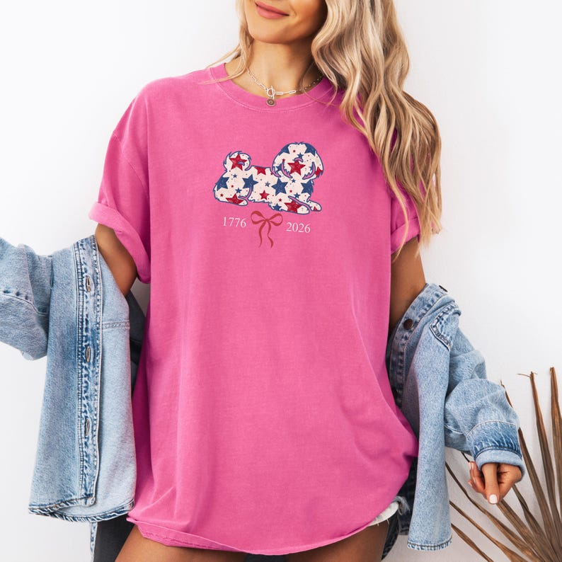 May include: A vibrant pink t-shirt with a dog design decorated with red and blue stars. The shirt displays the numbers "1776" and "2026" with a bow graphic. The sleeves are rolled up.