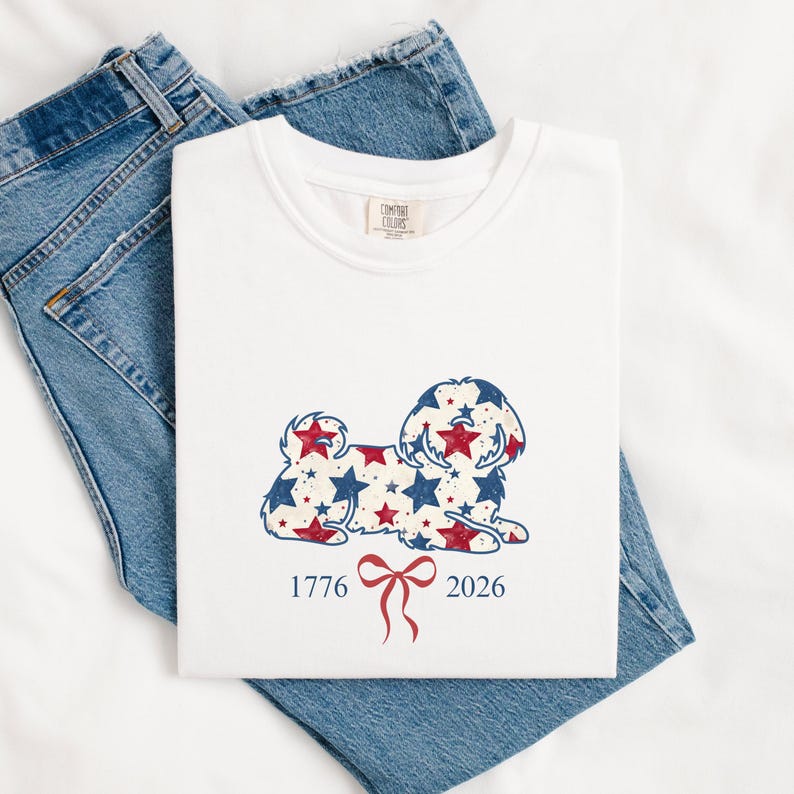 May include: White t-shirt featuring a dog design adorned with red and blue stars. The shirt displays the numbers "1776" and "2026" with a red bow graphic. The t-shirt is folded on a pair of blue denim jeans.