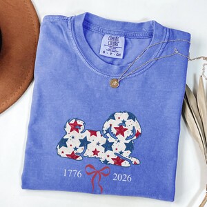 May include: A blue Comfort Colors t-shirt with a dog design made of red and blue stars. The shirt displays the numbers "1776" and "2026" with a red bow. A gold necklace is draped across the shirt.