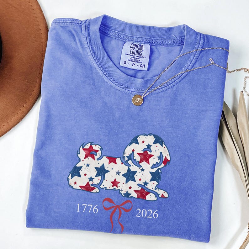 May include: A blue Comfort Colors t-shirt with a dog design made of red and blue stars. The shirt displays the numbers "1776" and "2026" with a red bow. A gold necklace is draped across the shirt.