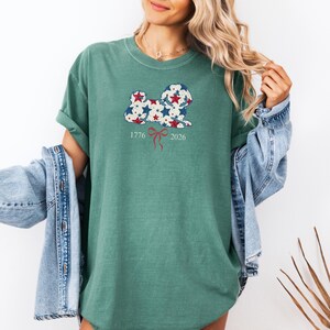 May include: A green t-shirt with a dog-shaped design filled with red and blue stars. The shirt has the numbers "1776" and "2026" with a red bow graphic. The t-shirt is made of a soft material.
