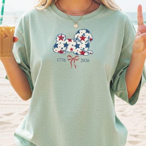 May include: A pale green t-shirt with a dog design filled with red and blue stars. Beneath the dog is a red bow and the numbers "1776" and "2026". The shirt has short sleeves and a loose fit.