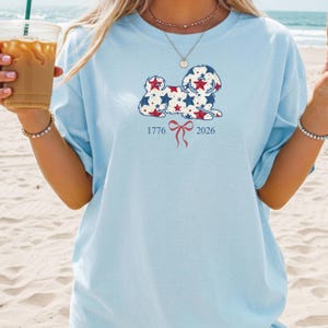 May include: Pale blue t-shirt with a dog design filled with red and blue stars. The shirt has the numbers "1776" and "2026" with a red bow graphic. The t-shirt is a relaxed, casual style.