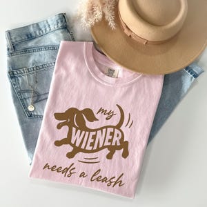 May include: A light pink t-shirt with the phrase "my wiener needs a leash" in brown lettering and a brown dachshund graphic. The shirt is paired with light blue jeans, a tan hat, and a necklace.