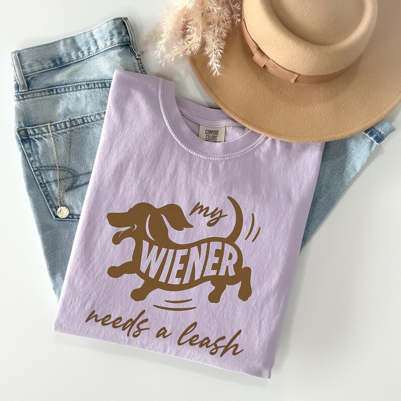 May include: A lilac-colored t-shirt with a brown dachshund graphic and the text "my wiener needs a leash." The shirt is paired with blue jeans, a tan hat, and a gold necklace.