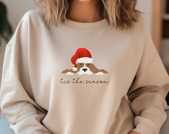 May include: Beige sweatshirt with a Basset Hound wearing a Santa hat graphic. The text "tis the season" is printed below the dog in a cursive font. The sweatshirt is a neutral colour, suitable for the festive season.