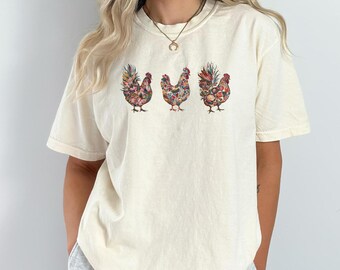 May include: A white t-shirt with a floral pattern of three chickens on the front. The chickens are in a row and are facing forward. The shirt is made of a soft, comfortable material and has a relaxed fit.